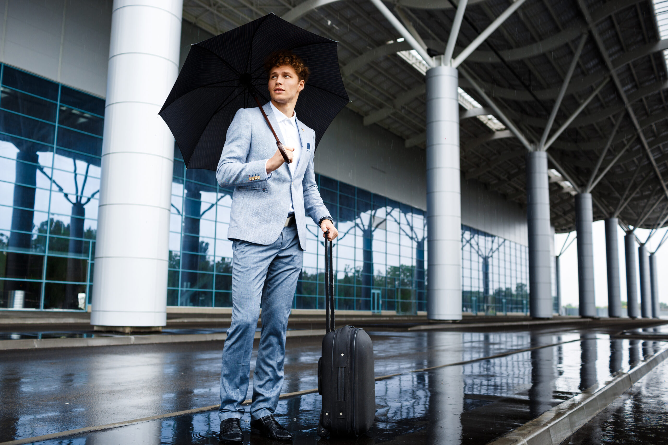 Picture of  young redhaired businessman looking aside holding black umbrella and suitcase in rain